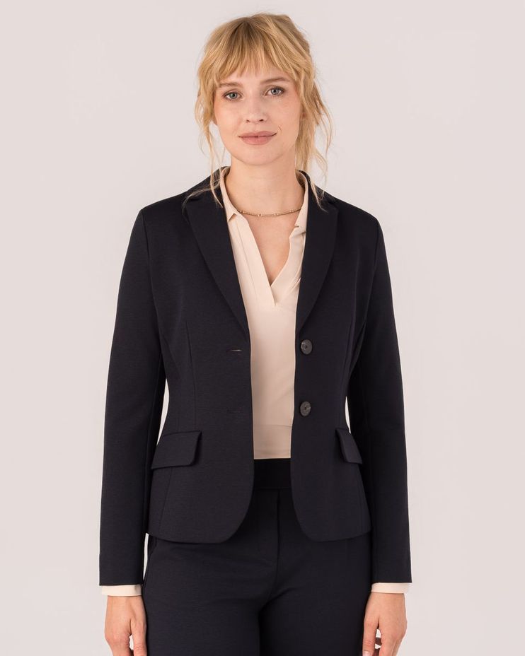 Frau im dunklen Blazer mit beige Bluse und Hose, professionell gekleidet, lächelnd.