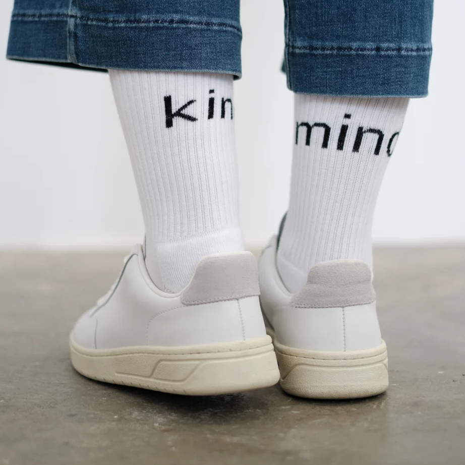Weiße Sneakers mit weißen Socken, auf denen „kinmino“ gedruckt ist, zu Jeans getragen.