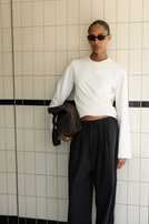 Person trägt oversized weißes Shirt, weite schwarze Hose und Sonnenbrille, hält Tasche, vor tesseliertem Hintergrund.