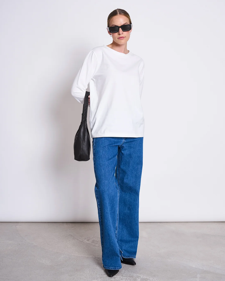 Frau trägt weißes oversized Shirt, blaue weite Hosen und schwarze Sonnenbrille mit Handtasche.